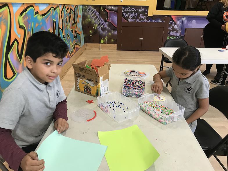 A boy folds a paper airplane as a girl makes art out of colored beads