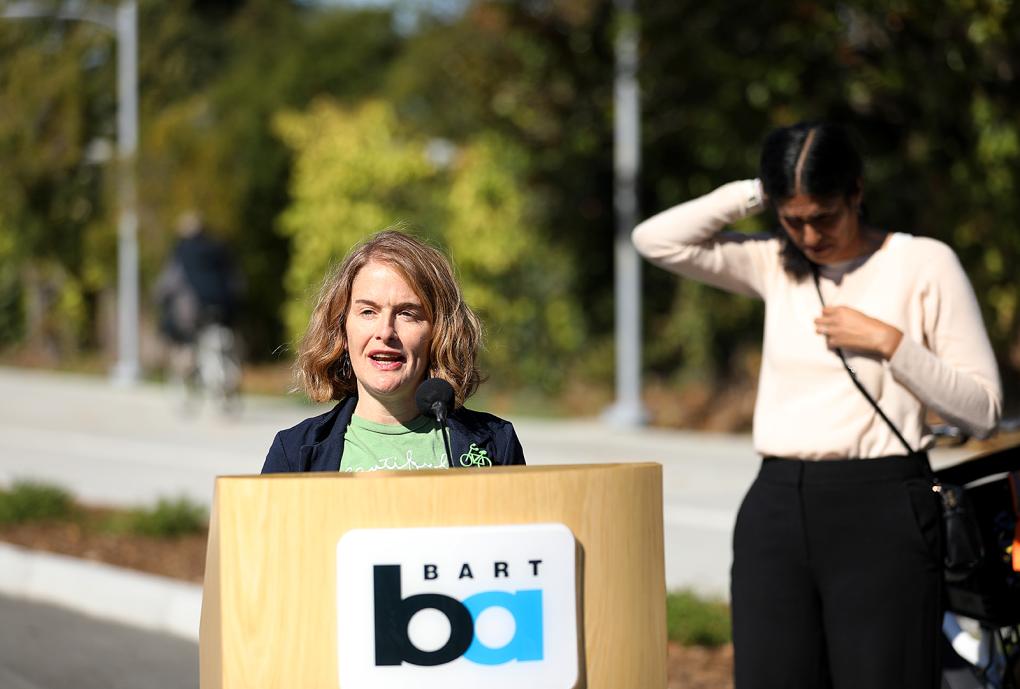 Co-executive director of Bike East Bay Rebecca Saltzman speaks at the ribbon cutting at a podium with the BART logo