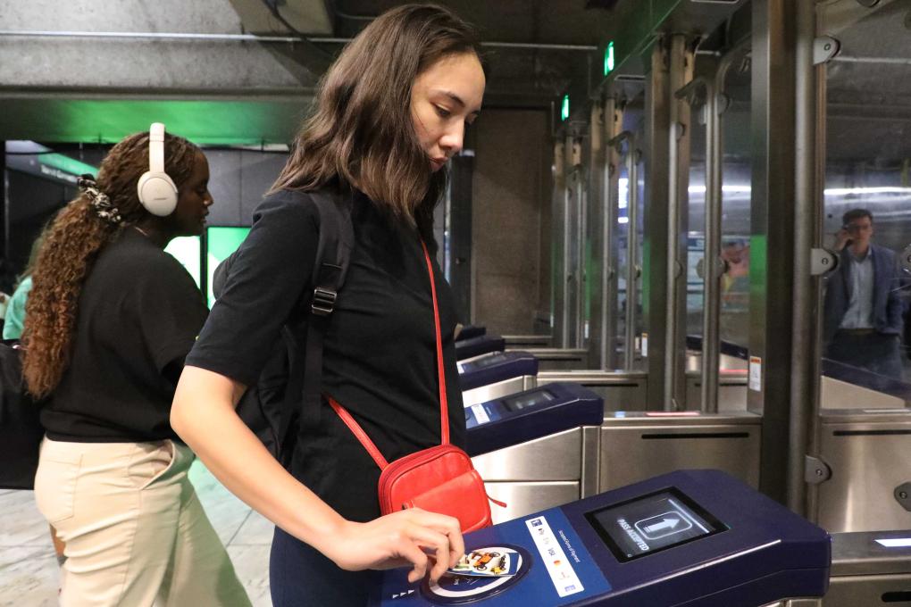 Person tapping a bank card at a BART fare gate at a station. A person is depicted in the background wearing headphones.