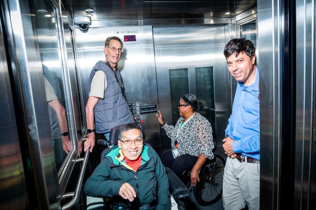 Four people pictured in a silver elevator, two men are standing, one man is seated in a wheelchair, and one woman is seated in a wheelchair pressing the elevator button