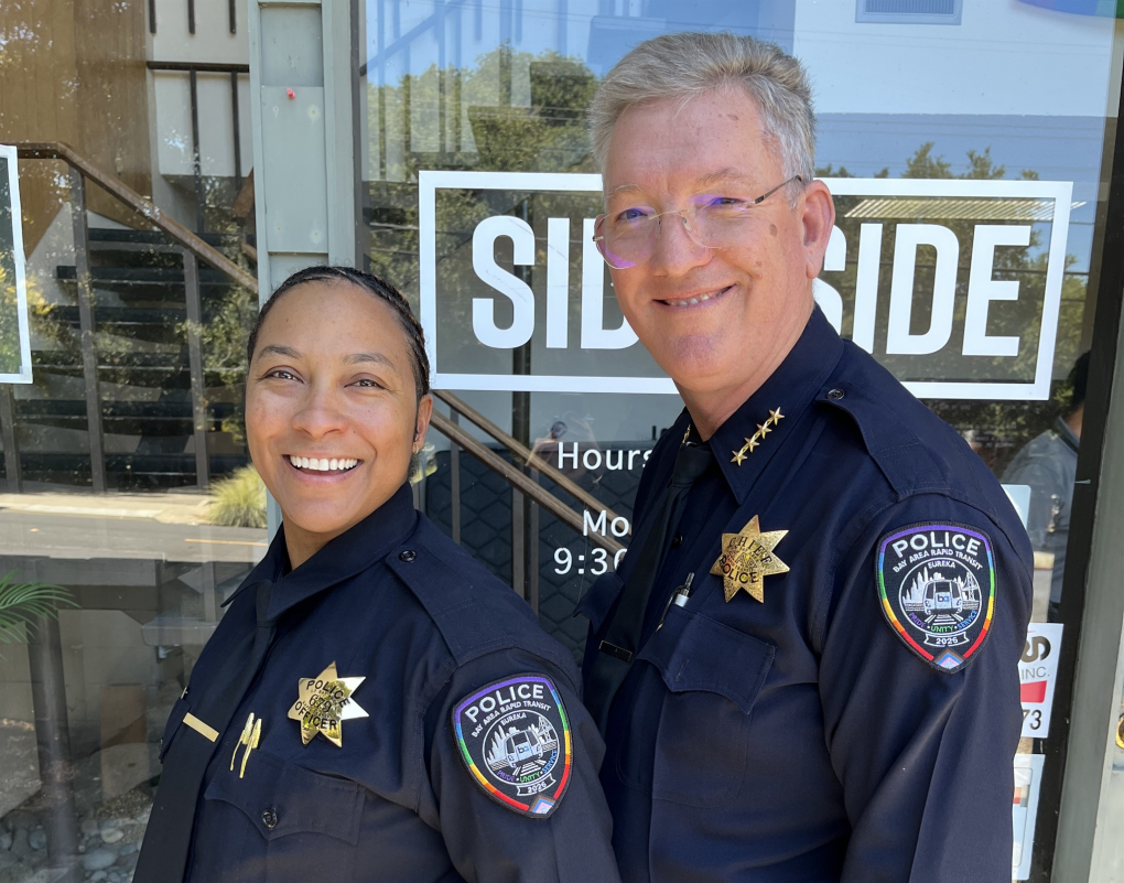 Officer Holmes and Chief Franklin show off their BART Police Pride Month patches in front of Side by Side.