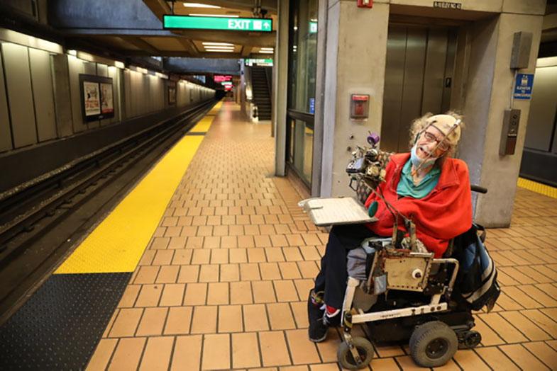 Hale Zukas is pictured on a BART platform in his wheelchair with a red sweatshirt and blue shirt in front of an elevator. The floor tiles are brown and yellow strip visible, as well as an exit sign. 