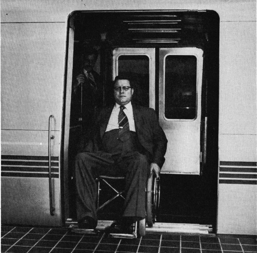A black and white photo of Harold Willson in a suit seated i his wheelchair on a train with the doors open on the platform.